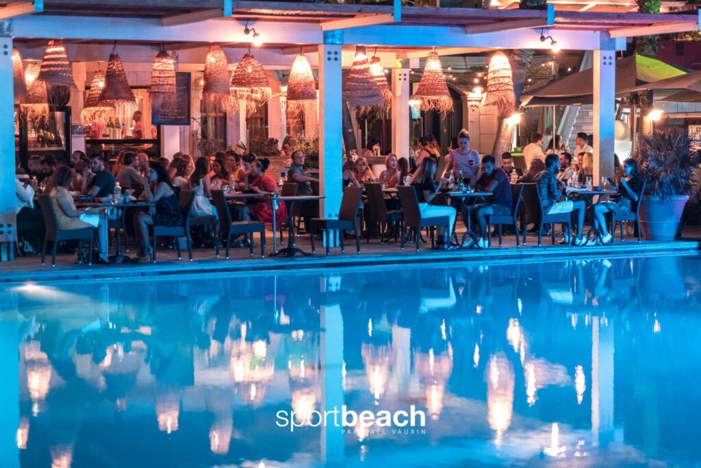 Restaurant extérieur éclairé le soir, convives aux tables en bois près de la piscine, suspensions en osier sous une structure blanche.