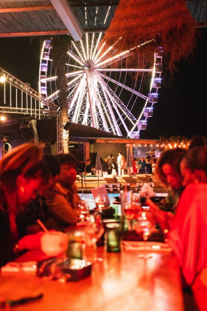 Personnes assises à une table en extérieur la nuit avec une grande roue lumineuse en arrière-plan.