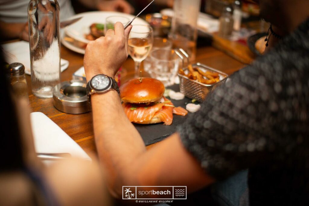 Personne tenant un couteau au-dessus d'un burger au saumon avec frites dans un petit panier sur une table en bois.